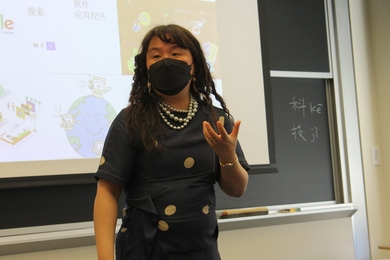 A woman in a polka-dot dress gestures while standing in front of a projection screen. 