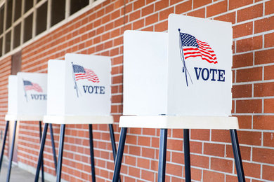 Photo shows 3 white voting boxes against a brick wall. The boxes have an American flag and say, “VOTE.”