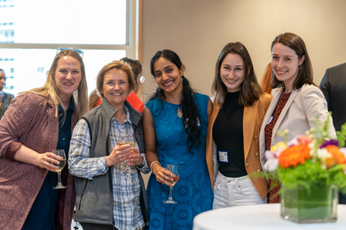 Five women stand together, posing for the camera in a crowded room