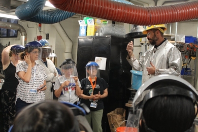 A man in a hard hat and mylar jacket lectures five teachers in a laboratory, all wearing hard hats with face shields