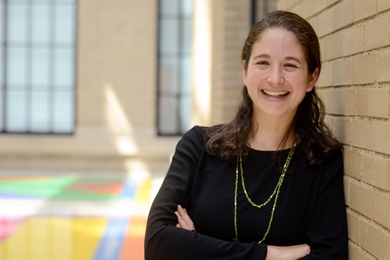 Elsa Olivetti leans against tan brick wall; windows and a colorful floor are in the background