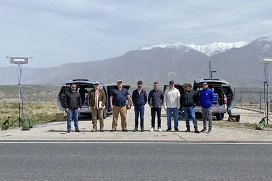 Photo of eight people standing in a line along a paved road on a sunny day. They stand in front of two vehicles with the trunks open and wires connected to small antennas on either side of the group. There are snow-capped mountains in the backround. 