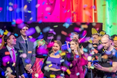 A shower of colorful confetti rains down on a group of people standing on stage. A young woman in the center is smiling and celebrating with her hands raised