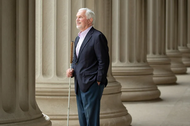 Paul Parravano stands among the columns of an iconic building on MIT’s campus, looking into the distance. He’s holding his white cane.