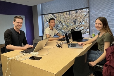 Three people sit around a table with laptops in front of a whiteboard and window 