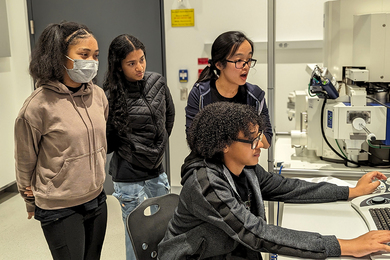Rebecca Li helps three middle schoolers, two standing and one seated at a computer, controlling a scanning electron microscope.
