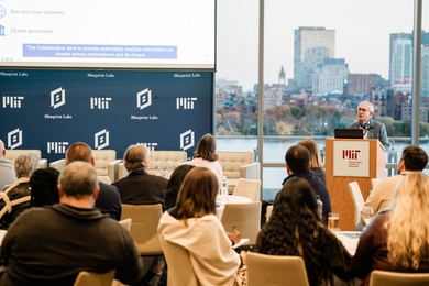 Josh Angrist speaks at a lectern, with the Charles River visible through a window behind him.