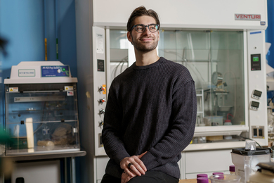 Branden Spitzer smiles while setting on a table inside a lab, with large equipment in background.