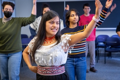 Five people stand in two rows indoors, all raising their left arm in unison. Soledad Chango, in the front, is wearing a traditional Ecuadorian dress.