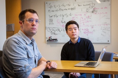 Two men sitting at a table, with a whiteboard in the background. 