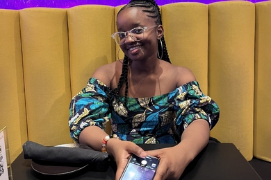 A young Black woman wearing a brightly patterned top and braids smiles over a set table at a restaurant. 