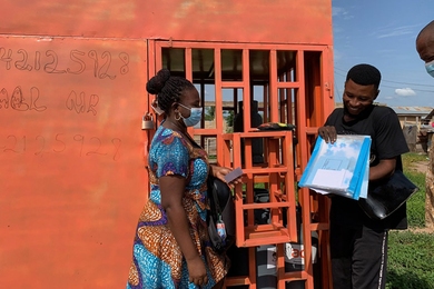 Standing outdoors in front of a large orange structure, a man shows a woman a square, handheld stitching machine.