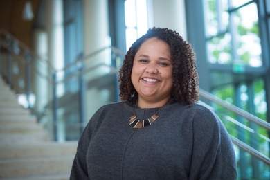 Portrait photo of Reimi Hicks standing next to an indoor stairway abutting a wall of windows