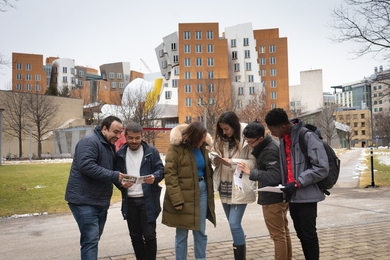 Six students wearing winter jackets consult printed materials and talk with each other in front of the Stata Center, a building of brick and steel with unusual angles