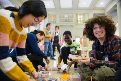 Four MIT sophomores crouch on the floor, smiling and building a structure using pencils, foam, and a ruler while two mentors look on.