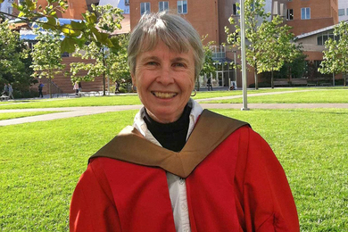 Mary-Lou Pardue, wearing a red academic robe, poses for a photo in front of a grassy MIT courtyard