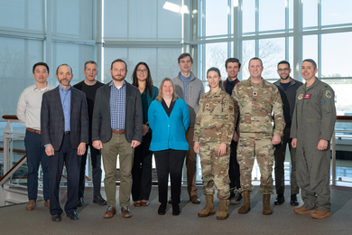 Twelve people, including three in military field uniforms, pose for a group photo in the Lincoln Laboratory main lobby, with a glass wall behind them