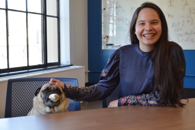 Ananda Santos Figueiredo seated at a table in a classroom, with a pug sitting in the chair next to her.