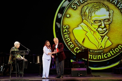 A seated woman and a standing man present an award to a young woman on stage. Behind them is a giant replica of the medal featuring a portrait of Stephen Hawking.