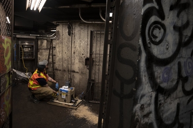 Kevin Geisel in orange safety vest is placing sampling equipment on the floor in a dark subway platform.