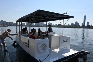 A man pushes a small, square solar boat with several passengers away from a dock. Boston’s skyline is the background.