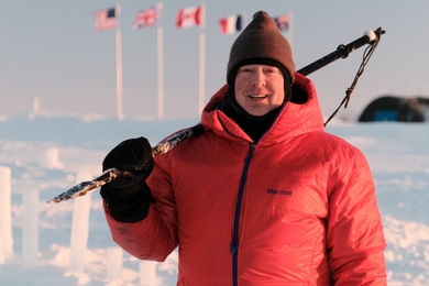 David Whelihan, wearing a red parka, stands on ice in the Arctic with a walking stick over his shoulder; flags representing different nations are pictured in the background.