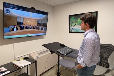 A graduate student stands in the Communication Studio, pitching to a virtual audience on a mounted screen.