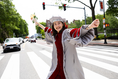 An MIT graduate in PhD graduation regalia smiles brightly as she crosses the street and holds a bouquet of flowers.