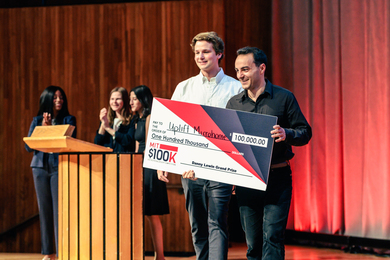 Trevor O’Leary and Charlie Nitschelm are on stage holding a giant $100K check awarded to “Uplight Microhome.”