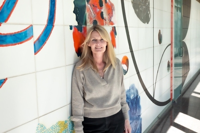 Karen O’Leary stands against a painted tile wall in well-lit hallway