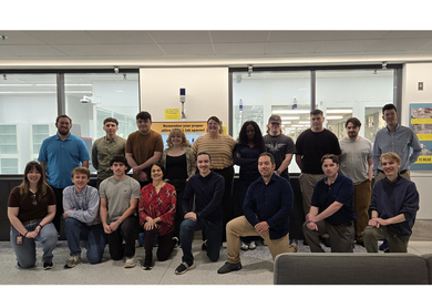 18 people pose in two rows in front of MIT.nano's cleanroom.