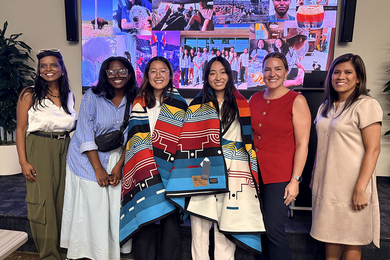 Six women pose standing in front of an electronic photo montage. Two in the center wear traditional Indigenous blankets.