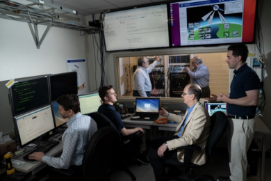 Six researchers are in a lab with computer screens, monitors, and two server racks of hardware 